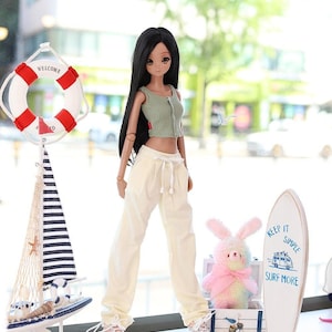 May include: A doll wearing a green crop top and cream-colored pants stands in front of a window. A decorative sailboat, a life preserver, and a surfboard with the text "KEEP IT SIMPLE SURF MORE" are also present. A pink bunny is also present.