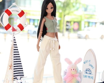 Puede incluir: Una muñeca con un top verde y pantalones color crema está de pie frente a una ventana. También hay un velero decorativo, un salvavidas y una tabla de surf con el texto "KEEP IT SIMPLE SURF MORE". También hay un conejito rosa.