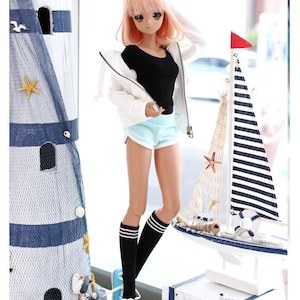May include: A doll with pink hair wearing a white hoodie, black tank top, light blue shorts, black and white striped knee-high socks, and blue sneakers. The doll is standing in front of a white lighthouse and a model sailboat.