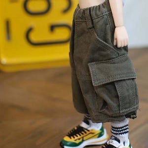 May include: Close-up of doll clothing: olive green cargo shorts with large pockets and a drawstring waist. The doll is wearing white socks with black stripes and yellow, green, and black sneakers.