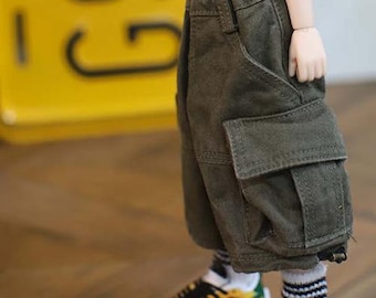 Puede incluir: Primer plano de ropa de muñeca: pantalones cortos cargo verde oliva con bolsillos grandes y cintura con cordón. La muñeca lleva calcetines blancos con rayas negras y zapatillas deportivas amarillas, verdes y negras.