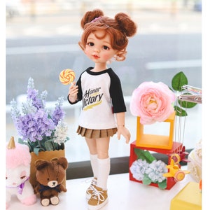 Puede incluir: Una mu&ntilde;eca con el pelo rojo en coletas que lleva una camiseta de b&eacute;isbol blanca y negra con las palabras "Honor Victory" impresas. La mu&ntilde;eca sostiene una piruleta amarilla y roja y lleva una falda plisada marr&oacute;n, calcetines blancos y zapatillas marrones. La mu&ntilde;eca est&aacute; de pie sobre una superficie blanca con una flor rosa, un oso de peluche marr&oacute;n y un peluche de unicornio blanco en el fondo.