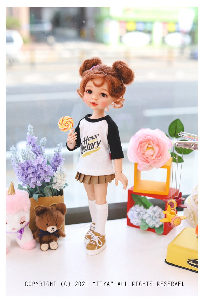 Puede incluir: Una mu&ntilde;eca con el pelo rojo en coletas que lleva una camiseta de b&eacute;isbol blanca y negra con las palabras "Honor Victory" impresas. La mu&ntilde;eca sostiene una piruleta amarilla y roja y lleva una falda plisada marr&oacute;n, calcetines blancos y zapatillas marrones. La mu&ntilde;eca est&aacute; de pie sobre una superficie blanca con una flor rosa, un oso de peluche marr&oacute;n y un peluche de unicornio blanco en el fondo.