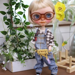 May include: A doll with blonde hair and brown-framed glasses, wearing a plaid shirt, denim overalls, and red and white sneakers. The doll has a yellow fanny pack with the text "TTYA". The doll is standing in front of greenery and flowers.
