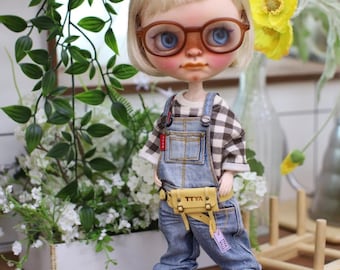 May include: A doll with blonde hair and brown-framed glasses, wearing a plaid shirt, denim overalls, and red and white sneakers. The doll has a yellow fanny pack with the text "TTYA". The doll is standing in front of greenery and flowers.