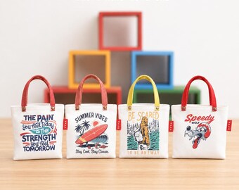 May include: Four white mini tote bags with red, yellow, and orange handles. Each bag features a unique graphic print: text reading "THE PAIN YOU FEEL TODAY IS THE STRENGTH YOU FEEL TOMORROW", a surfboard, a person in a hoodie, and a cartoon wolf.