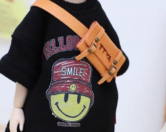 Puede incluir: Una sudadera negra con la palabra "ELLO" y un gráfico de una cara sonriente con la palabra "SMILE". Un pequeño bolso bandolera de cuero marrón claro con las letras "TTVA" se lleva sobre el hombro.