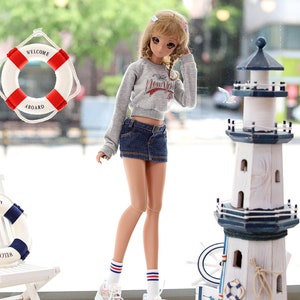 May include: A doll wearing a grey sweatshirt with "New York" printed on it, blue denim shorts, white socks with red and blue stripes, and pink and blue sneakers. The doll is standing in front of a white wooden lighthouse with a blue base.