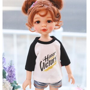 Puede incluir: Una mu&ntilde;eca con el pelo rojo en coletas que lleva una camiseta de b&eacute;isbol blanca y negra con las palabras "Honor y Victoria" impresas. La mu&ntilde;eca lleva pantalones cortos de mezclilla y calcetines blancos.