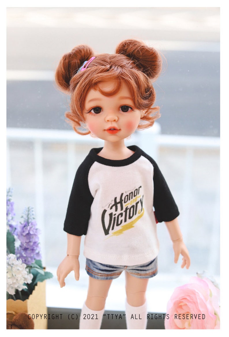 Puede incluir: Una mu&ntilde;eca con el pelo rojo en coletas que lleva una camiseta de b&eacute;isbol blanca y negra con las palabras "Honor y Victoria" impresas. La mu&ntilde;eca lleva pantalones cortos de mezclilla y calcetines blancos.