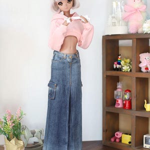 Puede incluir: Una mu&ntilde;eca con una sudadera rosa con capucha recortada y una falda vaquera azul con un bolsillo cargo grande. La mu&ntilde;eca est&aacute; de pie sobre un suelo de madera con un peque&ntilde;o estante de madera en el fondo.