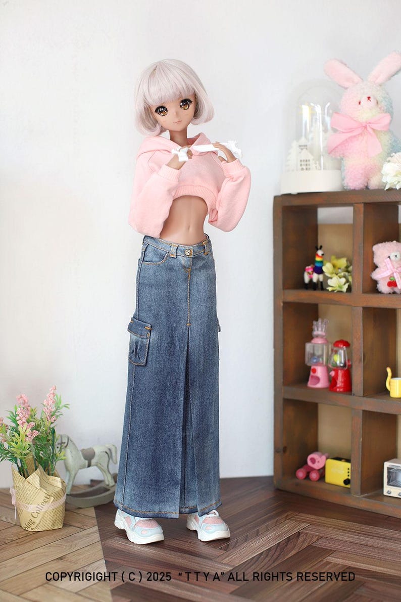 Puede incluir: Una mu&ntilde;eca con una sudadera rosa con capucha recortada y una falda vaquera azul con un bolsillo cargo grande. La mu&ntilde;eca est&aacute; de pie sobre un suelo de madera con un peque&ntilde;o estante de madera en el fondo.