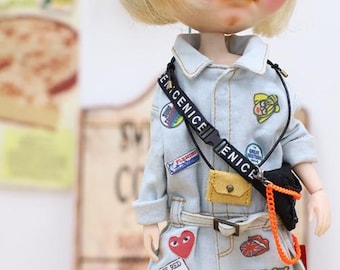 Puede incluir: Una muñeca con un mono vaquero azul claro adornado con varios parches y el texto "WAKE UP!". La muñeca tiene el pelo rubio, gafas naranjas y un pequeño bolso amarillo. Una correa negra en el pecho dice "ENICE".