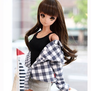 Puede incluir: Una mu&ntilde;eca con el pelo largo y casta&ntilde;o que lleva un top negro de tirantes, una camisa a cuadros azul y blanca y una falda vaquera gris. La mu&ntilde;eca est&aacute; de pie sobre una superficie de madera con un fondo borroso.