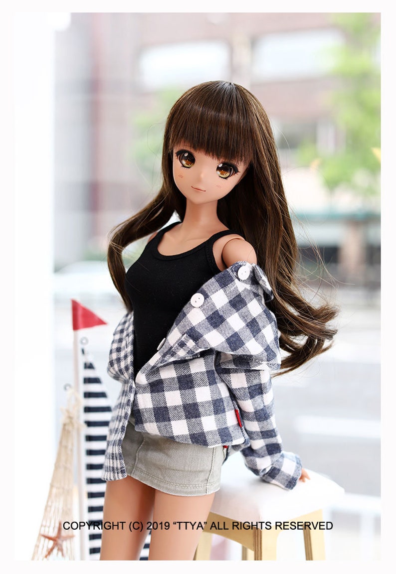 Puede incluir: Una mu&ntilde;eca con el pelo largo y casta&ntilde;o que lleva un top negro de tirantes, una camisa a cuadros azul y blanca y una falda vaquera gris. La mu&ntilde;eca est&aacute; de pie sobre una superficie de madera con un fondo borroso.