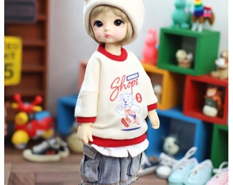 May include: A doll with blonde hair wearing a cream-colored beanie, a cream sweatshirt with red trim and a graphic of a dog running with the text "Shops", blue denim cargo shorts, white socks with red and blue stripes, and white lace-up sneakers.