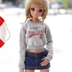 May include: A doll wearing a grey cropped sweatshirt with the text "AMERICAN COLLEGE LEAGUE NewYork" in red. The doll is also wearing a blue denim skirt and has blonde hair with a white and pink hair clip.