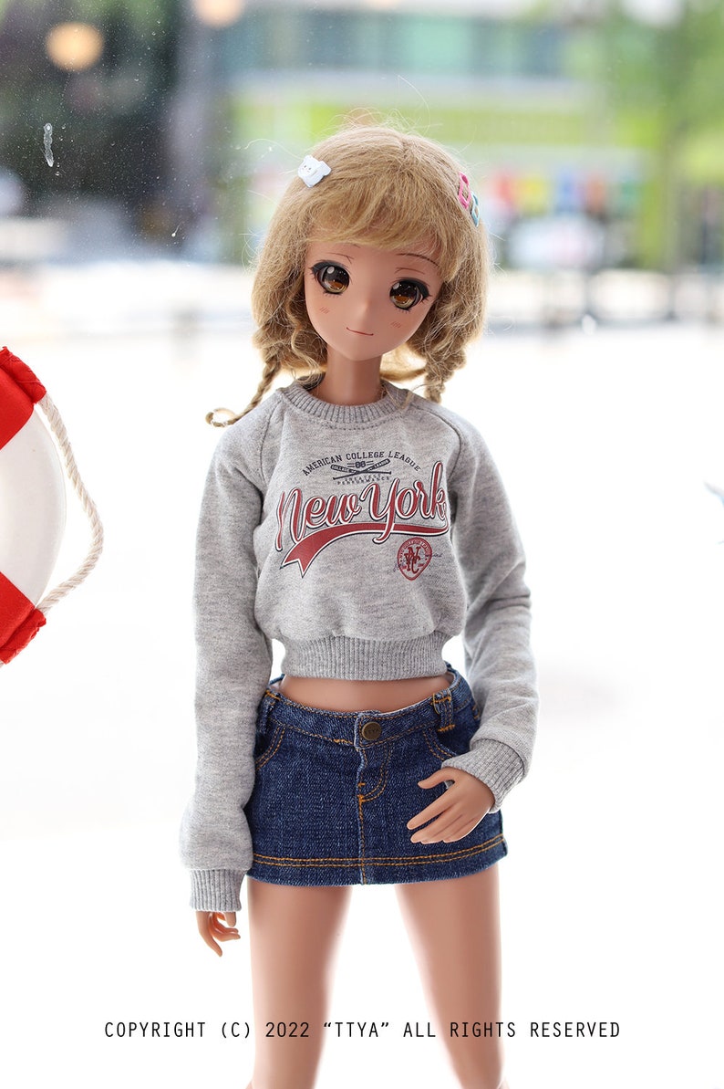 May include: A doll wearing a grey cropped sweatshirt with the text "AMERICAN COLLEGE LEAGUE NewYork" in red. The doll is also wearing a blue denim skirt and has blonde hair with a white and pink hair clip.