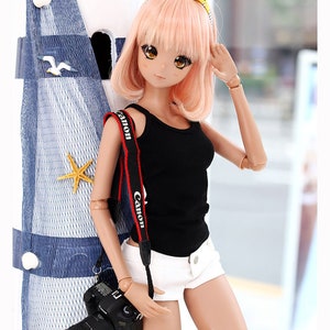 Puede incluir: Una mu&ntilde;eca con el pelo rosa que lleva un top negro sin mangas, pantalones cortos blancos y una diadema amarilla y blanca. La mu&ntilde;eca sostiene una c&aacute;mara negra con una correa roja de Canon.