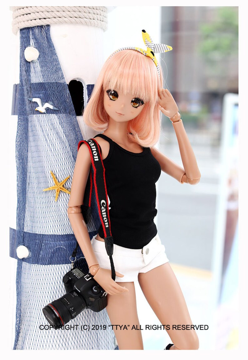 Puede incluir: Una mu&ntilde;eca con el pelo rosa que lleva un top negro sin mangas, pantalones cortos blancos y una diadema amarilla y blanca. La mu&ntilde;eca sostiene una c&aacute;mara negra con una correa roja de Canon.