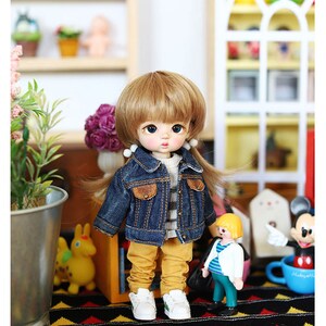 以下が含まれることがあります： 金髪のウィッグをつけた人形が、青いデニムジャケット、ストライプのシャツ、黄色のズボンを着用しています。人形は黒と黄色の模様の敷物の上に立っており、背景には他の玩具があります。