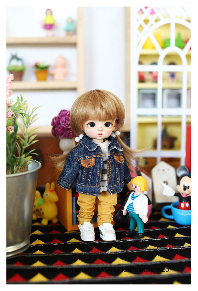 以下が含まれることがあります： 金髪のウィッグをつけた人形が、青いデニムジャケット、ストライプのシャツ、黄色のズボンを着用しています。人形は黒と黄色の模様の敷物の上に立っており、背景には他の玩具があります。