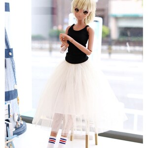 K&ouml;nnte beinhalten: Eine Puppe tr&auml;gt einen wei&szlig;en T&uuml;llrock, ein schwarzes Tanktop, wei&szlig;e Socken mit roten und blauen Streifen und blaue Turnschuhe. Die Puppe hat blonde Haare und braune Augen. Die Puppe steht vor einem Fenster.