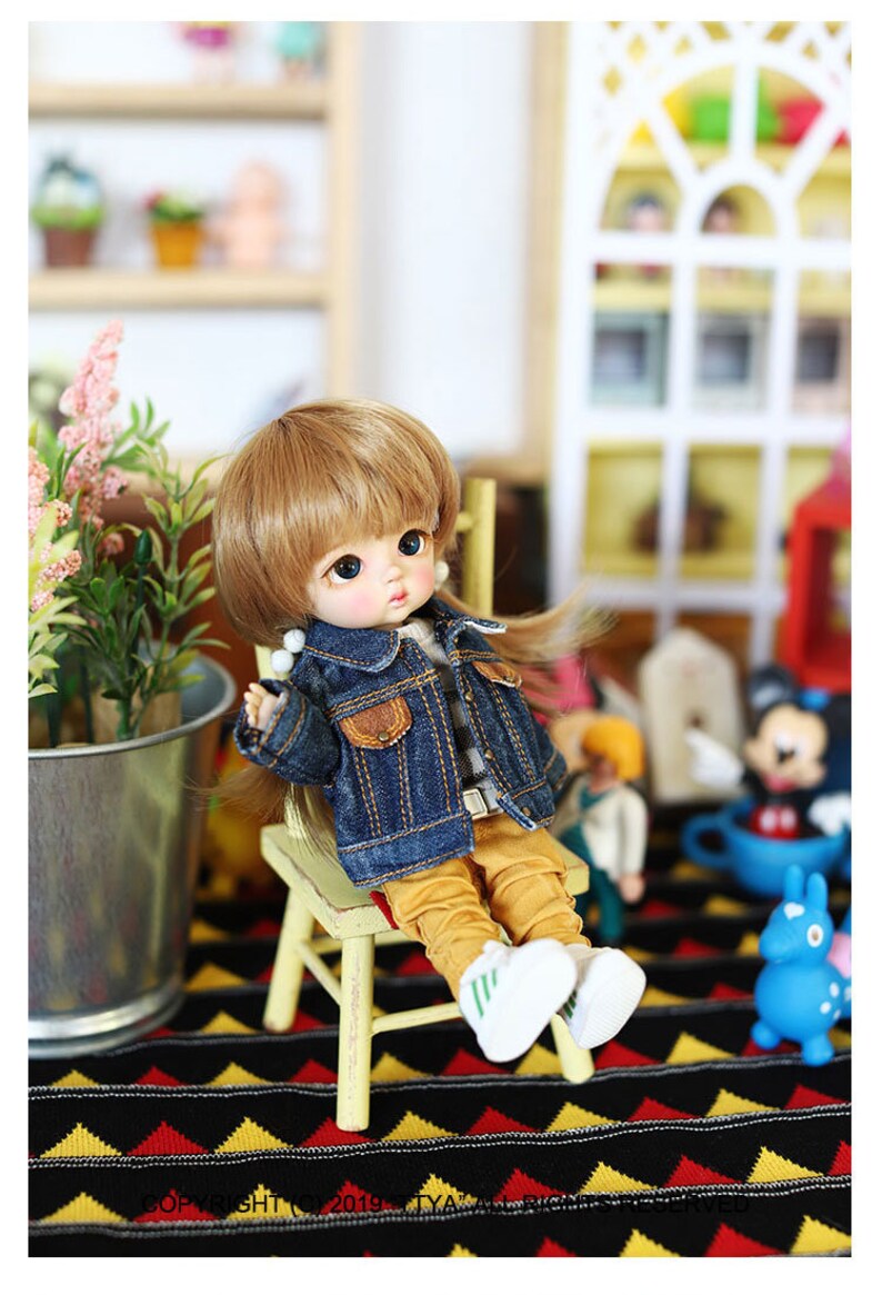 以下が含まれることがあります： 青いデニムジャケット、黄色のズボン、白いスニーカーを履いた小さな人形が木製の椅子に座っています。人形はブロンドの髪と青い目をしています。椅子は黒と黄色の模様の敷物の上にあります。
