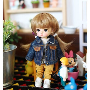 以下が含まれることがあります： 金髪の小さな人形が、青いデニムジャケット、ストライプのシャツ、黄色のズボンを着ています。人形は、黒と黄色の模様の敷物の上に立っており、背景には他の人形や玩具があります。