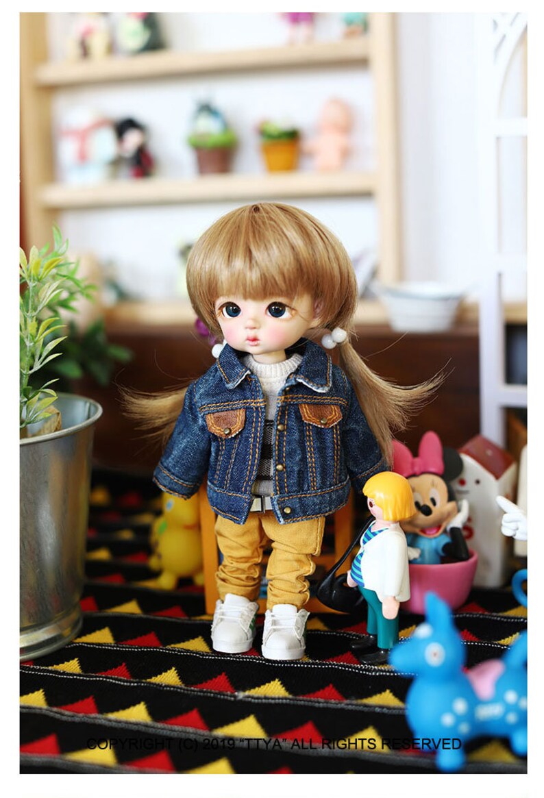 以下が含まれることがあります： 金髪の小さな人形が、青いデニムジャケット、ストライプのシャツ、黄色のズボンを着ています。人形は、黒と黄色の模様の敷物の上に立っており、背景には他の人形や玩具があります。