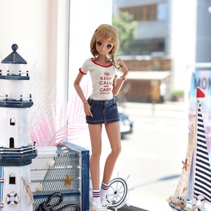 Puede incluir: Una mu&ntilde;eca con una camiseta blanca con texto rojo que dice "Keep Calm and Carry a Tiyado" y una falda vaquera azul. La mu&ntilde;eca est&aacute; de pie frente a una ventana con vista a una calle de la ciudad. La mu&ntilde;eca lleva calcetines blancos con rayas rojas y azules y zapatillas blancas. La mu&ntilde;eca tiene el pelo largo y casta&ntilde;o y ojos azules.