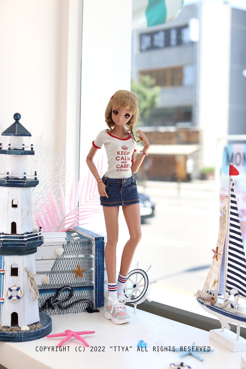 Puede incluir: Una mu&ntilde;eca con una camiseta blanca con texto rojo que dice "Keep Calm and Carry a Tiyado" y una falda vaquera azul. La mu&ntilde;eca est&aacute; de pie frente a una ventana con vista a una calle de la ciudad. La mu&ntilde;eca lleva calcetines blancos con rayas rojas y azules y zapatillas blancas. La mu&ntilde;eca tiene el pelo largo y casta&ntilde;o y ojos azules.