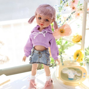 Pode incluir: Uma boneca com cabelo rosa usando um moletom com capuz lavanda e uma saia jeans. A boneca est&aacute; em p&eacute; em uma superf&iacute;cie branca com uma pequena televis&atilde;o atr&aacute;s dela. A televis&atilde;o tem um animal de pel&uacute;cia branco dentro. H&aacute; flores no fundo.