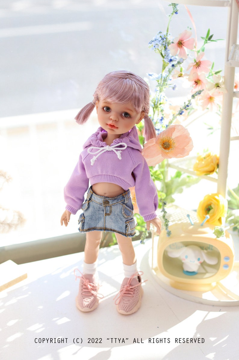 Pode incluir: Uma boneca com cabelo rosa usando um moletom com capuz lavanda e uma saia jeans. A boneca est&aacute; em p&eacute; em uma superf&iacute;cie branca com uma pequena televis&atilde;o atr&aacute;s dela. A televis&atilde;o tem um animal de pel&uacute;cia branco dentro. H&aacute; flores no fundo.