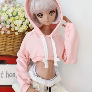 Puede incluir: Una sudadera con capucha rosa corta con cordones blancos y mangas largas. La sudadera la lleva una mu&ntilde;eca con el pelo largo y blanco y los ojos marrones.