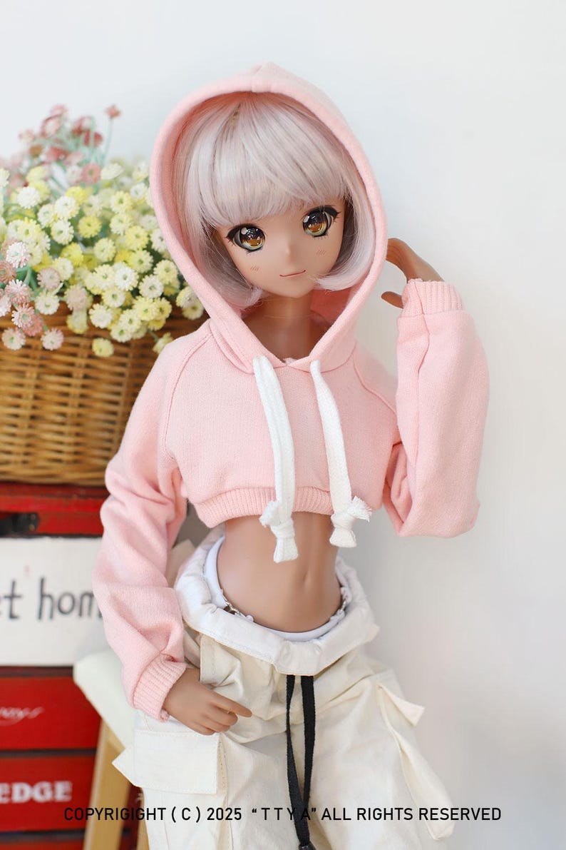 Puede incluir: Una sudadera con capucha rosa corta con cordones blancos y mangas largas. La sudadera la lleva una mu&ntilde;eca con el pelo largo y blanco y los ojos marrones.