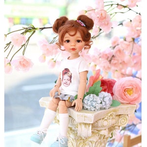 Puede incluir: Una mu&ntilde;eca con el pelo casta&ntilde;o en coletas que lleva una camiseta blanca con una imagen impresa de una mu&ntilde;eca, una falda azul y blanca, calcetines blancos y zapatillas azules. La mu&ntilde;eca est&aacute; sentada en una columna blanca con flores rosas y azules alrededor. El texto "COPYRIGHT (C) 2021 "TTYA" ALL RIGHTS RESERVED" est&aacute; en la parte inferior de la imagen.