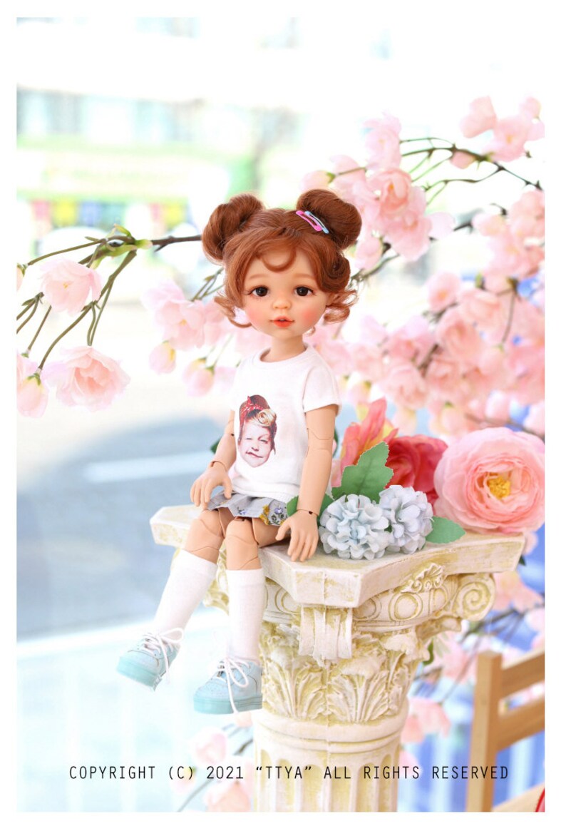 Puede incluir: Una mu&ntilde;eca con el pelo casta&ntilde;o en coletas que lleva una camiseta blanca con una imagen impresa de una mu&ntilde;eca, una falda azul y blanca, calcetines blancos y zapatillas azules. La mu&ntilde;eca est&aacute; sentada en una columna blanca con flores rosas y azules alrededor. El texto "COPYRIGHT (C) 2021 "TTYA" ALL RIGHTS RESERVED" est&aacute; en la parte inferior de la imagen.