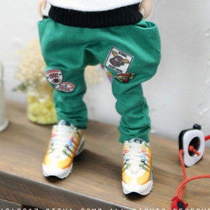 May include: A doll wearing a white shirt with a red and black design, green pants with patches, and colorful sneakers.