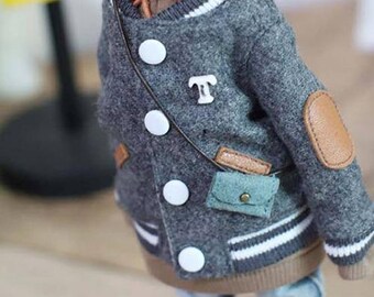 Puede incluir: Una chaqueta gris para muñeca con rayas blancas y botones blancos grandes. La chaqueta tiene un pequeño parche de cuero en el codo y un pequeño bolso turquesa. Una letra "T" blanca está en el pecho.