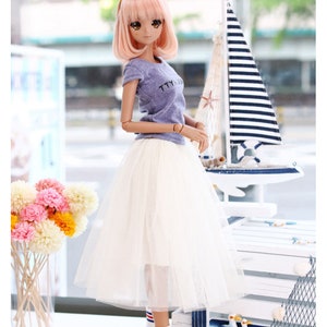 K&ouml;nnte beinhalten: Eine Puppe tr&auml;gt ein hellblaues T-Shirt mit dem Text "TTYL" darauf, einen wei&szlig;en T&uuml;llrock, blau-wei&szlig; gestreifte Socken und blaue Turnschuhe. Die Puppe hat rosa Haare und tr&auml;gt ein gelbes Stirnband. Die Puppe steht vor einer wei&szlig;en Holzkiste mit nautischem Thema.