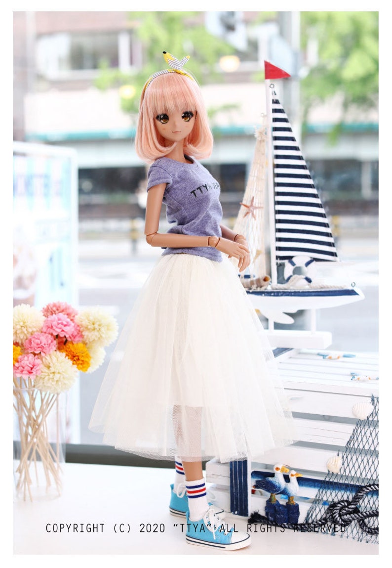 K&ouml;nnte beinhalten: Eine Puppe tr&auml;gt ein hellblaues T-Shirt mit dem Text "TTYL" darauf, einen wei&szlig;en T&uuml;llrock, blau-wei&szlig; gestreifte Socken und blaue Turnschuhe. Die Puppe hat rosa Haare und tr&auml;gt ein gelbes Stirnband. Die Puppe steht vor einer wei&szlig;en Holzkiste mit nautischem Thema.