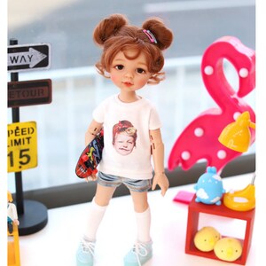 Puede incluir: Una mu&ntilde;eca con el pelo casta&ntilde;o recogido en dos mo&ntilde;os, lleva una camiseta blanca con una imagen impresa de una persona, pantalones cortos de mezclilla azul, calcetines blancos y zapatillas de deporte azul claro. La mu&ntilde;eca sostiene una peque&ntilde;a patineta en una mano. La mu&ntilde;eca est&aacute; de pie sobre una superficie blanca frente a una l&aacute;mpara en forma de flamenco rosa.