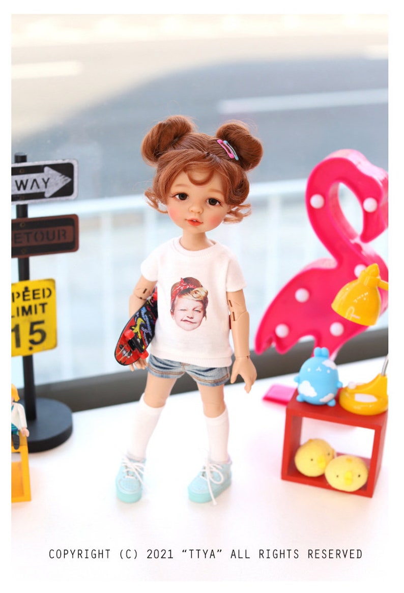 Puede incluir: Una mu&ntilde;eca con el pelo casta&ntilde;o recogido en dos mo&ntilde;os, lleva una camiseta blanca con una imagen impresa de una persona, pantalones cortos de mezclilla azul, calcetines blancos y zapatillas de deporte azul claro. La mu&ntilde;eca sostiene una peque&ntilde;a patineta en una mano. La mu&ntilde;eca est&aacute; de pie sobre una superficie blanca frente a una l&aacute;mpara en forma de flamenco rosa.