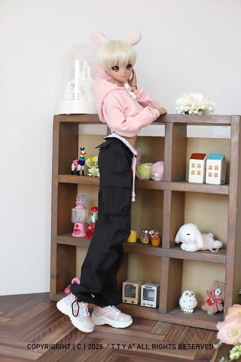 Puede incluir: Una mu&ntilde;eca con una sudadera rosa, pantalones cargo negros y zapatillas blancas. La mu&ntilde;eca est&aacute; de pie junto a una estanter&iacute;a de madera con varios art&iacute;culos decorativos.