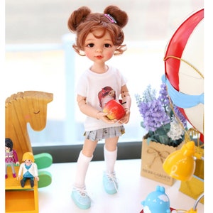 Puede incluir: Una mu&ntilde;eca con el pelo casta&ntilde;o en coletas que lleva una camiseta blanca con un gr&aacute;fico de una ni&ntilde;a, una falda gris, calcetines blancos y zapatos azules. La mu&ntilde;eca est&aacute; sosteniendo una manzana roja. La mu&ntilde;eca est&aacute; de pie sobre una superficie blanca con un caballo de balanc&iacute;n de madera, una peque&ntilde;a l&aacute;mpara y un globo aerost&aacute;tico en el fondo.