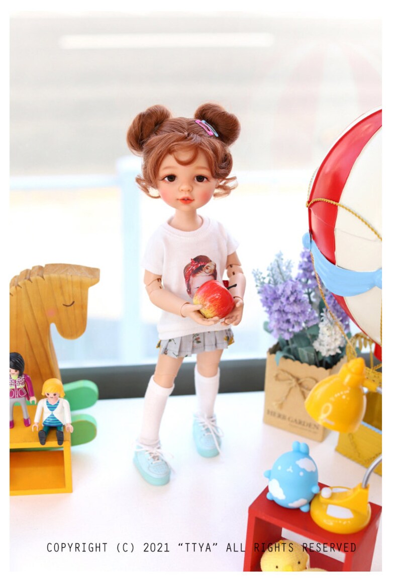 Puede incluir: Una mu&ntilde;eca con el pelo casta&ntilde;o en coletas que lleva una camiseta blanca con un gr&aacute;fico de una ni&ntilde;a, una falda gris, calcetines blancos y zapatos azules. La mu&ntilde;eca est&aacute; sosteniendo una manzana roja. La mu&ntilde;eca est&aacute; de pie sobre una superficie blanca con un caballo de balanc&iacute;n de madera, una peque&ntilde;a l&aacute;mpara y un globo aerost&aacute;tico en el fondo.