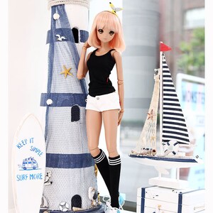 Puede incluir: Una mu&ntilde;eca con el pelo rosa que lleva un top negro sin mangas, pantalones cortos blancos, calcetines a rayas blancas y negras y zapatillas azules. La mu&ntilde;eca est&aacute; de pie junto a un faro blanco con rayas azules y blancas y una tabla de surf blanca con el texto "Keep it simple, surf more".