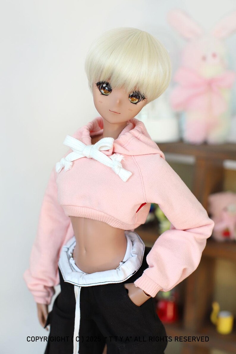 Puede incluir: Una sudadera con capucha rosa recortada con un cord&oacute;n blanco y un lazo blanco. La sudadera la lleva una mu&ntilde;eca con el pelo rubio y los ojos marrones.