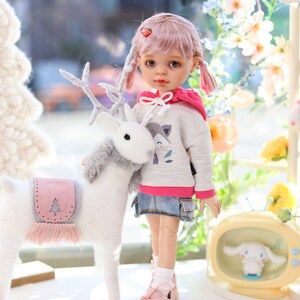 Pode incluir: Uma boneca rosa e cinza com cabelo rosa e tran&ccedil;as usando um moletom cinza com um gr&aacute;fico de raposa e uma saia jeans. A boneca est&aacute; segurando uma rena de pel&uacute;cia branca com chifres. A boneca est&aacute; usando t&ecirc;nis rosa.